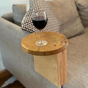 Op de afbeelding: Een houten bijzettafel met een rond blad en een rechthoekige basis. De tafel is bevestigd aan de zijkant van een bank en heeft een glas rode wijn erop.