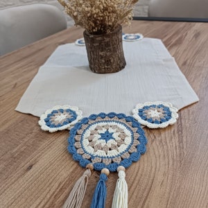May include: A crocheted table runner with three blue and beige crocheted coasters. The coasters have a star design in the center. The table runner is on a wooden table.