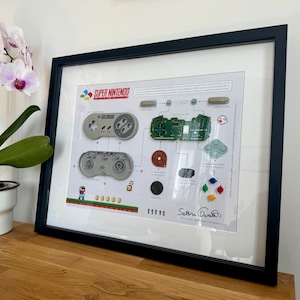Op de afbeelding: Een ingelijste print van een gedemonteerde Super Nintendo-controller met een diagram dat laat zien hoe de onderdelen in elkaar passen. De print bevat de tekst "Super Nintendo Entertainment System" en "Saturn Quartz".