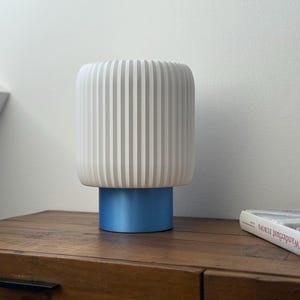 Può includere: Una lampada da tavolo bianca con una base blu. Il paralume ha scanalature verticali ed è realizzato in materiale bianco. La lampada è appoggiata su un tavolo di legno.