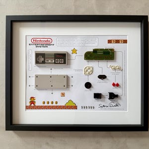 May include: A framed display of a disassembled Nintendo Entertainment System, featuring the console, controller, and various components. The display includes a small pixelated image of Mario from the Super Mario Bros. video game.