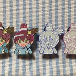 May include: Four enamel pins featuring a cartoon character with a surprised expression. The character is wearing a blue and white spacesuit and has a red and white light on top of their head. The pins are in different colour variations: gold, silver, purple, and light blue.