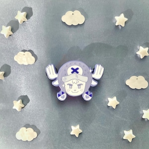 以下が含まれることがあります： 白い帽子と青いXがついた漫画のキャラクターが描かれた、紫と白のエナメルピン。キャラクターは青い目とピンクの口をした白い顔をしています。ピンは白い雲と星に囲まれています。