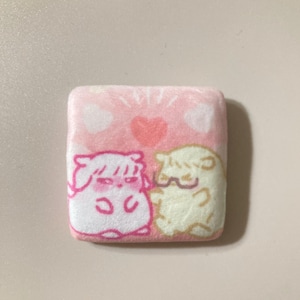 May include: A square, pink plush item with a cartoon illustration of two characters. The top of the item has a pink background with white hearts and a red heart in the center. The characters are in white and yellow with pink outlines.