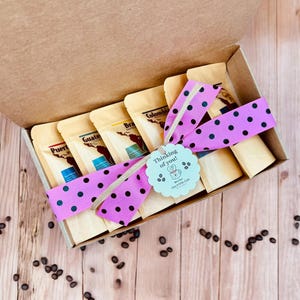 May include: A gift box filled with six small bags of coffee beans, tied with a pink polka dot ribbon and a tag that says "Thinking of you!" The box is open and sitting on a wooden surface with coffee beans scattered around it.
