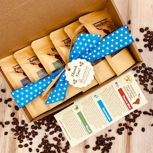 May include: A gift box filled with six small bags of coffee beans, each labeled with a different country of origin. The box is tied with a blue ribbon with white polka dots and a "Thank You!" tag. The box is surrounded by coffee beans.
