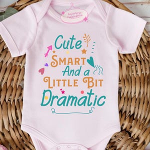 May include: A light pink baby bodysuit with the text "Cute Smart And a Little Bit Dramatic" in teal, orange, and purple. The bodysuit is short-sleeved and has a snap closure. The bodysuit is on a woven basket.