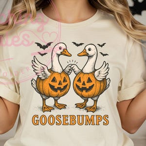 Peut inclure: T-shirt crème avec un motif d'Halloween. Le motif comprend deux oies avec des corps de citrouille, des ailes et des becs, ainsi que le mot "GOOSEBUMPS" en orange. Des chauves-souris noires sont également incluses dans le motif.