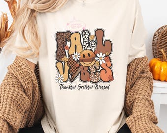 May include: Cream-colored t-shirt with the words "Fall Vibes" in a retro font, featuring a smiley face and floral accents. The text "Thankful Grateful Blessed" is below the main text. The shirt has a relaxed fit.