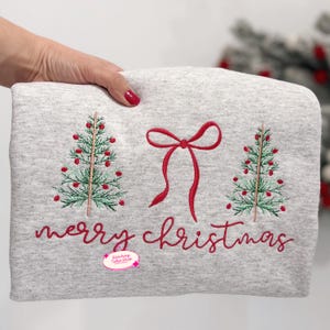 Könnte beinhalten: Ein hellgraues Sweatshirt mit gestickten Weihnachtsbäumen und einer roten Schleife. Die Worte "merry christmas" sind in roter Schrift gestickt. Das Sweatshirt wird von einer Hand mit rotem Nagellack gehalten.