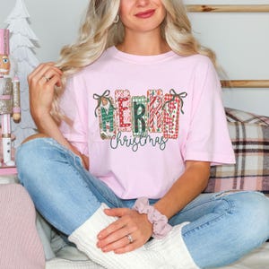 Può includere: Una maglietta rosa chiaro con la scritta "MERRY Christmas" in un design festivo. Le lettere sono decorate con motivi e fiocchi, creando un tema natalizio. La maglietta è realizzata in un materiale morbido.