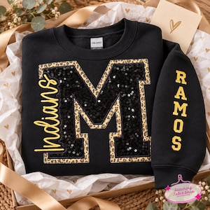 May include: A black sweatshirt featuring a large letter M filled with black sequins and outlined in a leopard print pattern. The word Indians is embroidered in yellow script on the left, and the name RAMOS is embroidered in yellow on the sleeve.