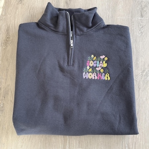 May include: A dark grey quarter-zip sweatshirt with a high collar. The sweatshirt has a colourful floral design and the words "SOCIAL WORKER" embroidered on the front. The zipper is silver.