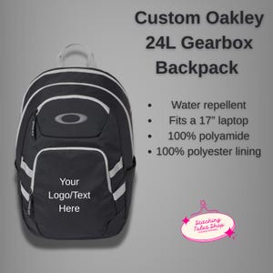 May include: Black Oakley 24L Gearbox backpack with white accents and a white handle. The backpack has a front pocket and a main compartment. The text "Your Logo/Text Here" is on the front. It fits a 17" laptop and is water repellent.