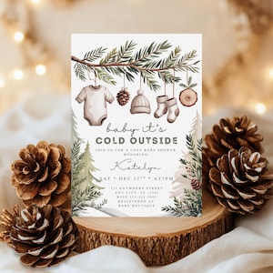May include: A baby shower invitation with a winter theme. The invitation features a watercolor illustration of baby clothes, a pinecone, a hat, and socks hanging from a branch. The text reads "baby it's cold outside" and includes event details.