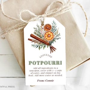 Op de afbeelding: Een cadeau verpakt in bruin papier, vastgebonden met touw, en een decoratief label. Het label toont een aquarel illustratie van gedroogde sinaasappelschijfjes, kaneelstokjes en groen. De tekst op het label luidt "STOVETOP POTPOURRI".