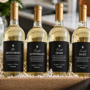 May include: Four glass wine bottles with gold foil caps and black labels in a woven basket. Each label features a small heart icon, custom text including The Honeymoon, First Anniversary, First Home, and First Fight, plus names and a date.