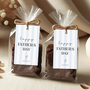 Puede incluir: Dos bolsas transparentes de celofán atadas con hilo, cada una con un brownie y una etiqueta. Las etiquetas tienen rayas azules y blancas y dicen "Happy Father's Day" con un corazón y el nombre "Sophie".