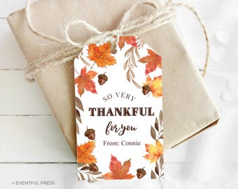 May include: A gift tag featuring watercolour illustrations of autumn leaves and acorns in shades of orange and red. The tag says "So Very Thankful for you" with the name "Connie" below. The tag is tied to a wrapped gift with twine.