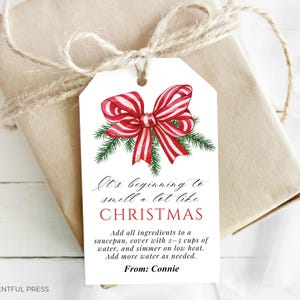 May include: A Christmas gift tag featuring a red and white striped bow and green foliage. The tag says "It's beginning to smell a lot like CHRISTMAS" with instructions and "From: Connie". The gift is wrapped in brown paper and tied with twine.