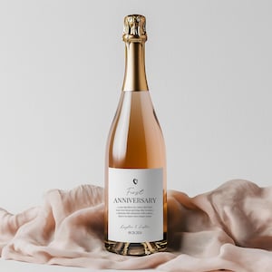 May include: A glass bottle of rose-colored sparkling wine with a gold foil neck wrap. The white label features a small heart icon, the text First ANNIVERSARY, a short message, the names Kaylee & Justin, and the date 09.28.2024 in black ink.