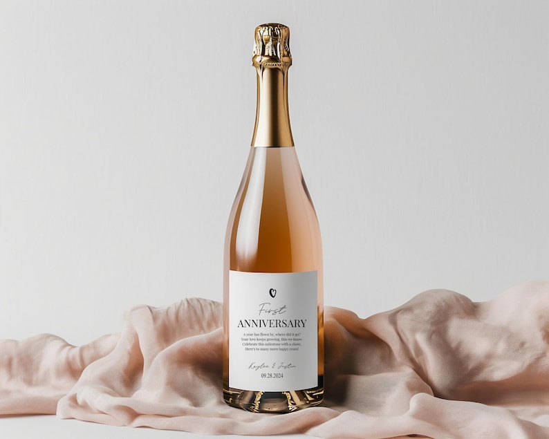 May include: A glass bottle of rose-colored sparkling wine with a gold foil neck wrap. The white label features a small heart icon, the text First ANNIVERSARY, a short message, the names Kaylee & Justin, and the date 09.28.2024 in black ink.