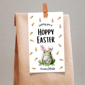 May include: A white Easter gift tag with the text "Wishing you a Hoppy Easter" and a green frog wearing a pink bunny ear hat. The tag is decorated with orange carrot illustrations and is clipped to a brown paper bag.