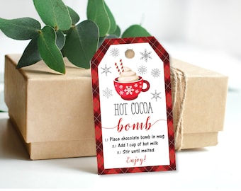 May include: A gift box tied with twine, adorned with a red and white plaid tag. The tag features a red mug with whipped cream and the words "HOT COCOA bomb". Instructions on the tag read: Place chocolate bomb in mug, add 1 cup of hot milk, stir until melted. Enjoy!