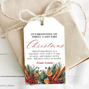 Könnte beinhalten: Ein Weihnachtsgeschenkanhänger mit dem Text "IT'S BEGINNING TO SMELL A LOT LIKE Christmas" in roter Schrift. Der Anhänger ist mit Tannenzweigen, Zimtstangen, getrockneten Orangenscheiben und Beeren verziert. Das Geschenk ist in braunes Papier gewickelt und mit Bindfaden verschnürt.