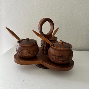 May include: A wooden spice rack with three small, round, lidded bowls, each with a small spoon. The bowls and spoons are a dark brown color. The rack has a handle and a decorative carved design.