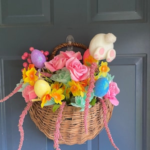 May include: A woven basket filled with pink roses, yellow daffodils, and decorative Easter eggs. A white bunny tail with pink paw pads peeks out from the arrangement. Pink, trailing stems add a whimsical touch to this Easter-themed decoration.