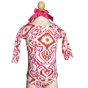 May include: A white long-sleeved bodysuit with a pink and red geometric pattern. The bodysuit has a scoop neck and is on a mannequin with a pink bow.
