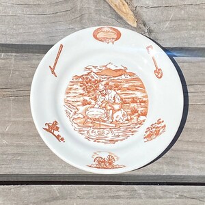 Peut inclure: Une assiette en céramique blanche avec une illustration orange détaillée d'un chercheur d'or. L'assiette présente une bordure avec des outils miniers et d'autres images connexes. Une pièce décorative pour une cuisine ou une salle à manger.