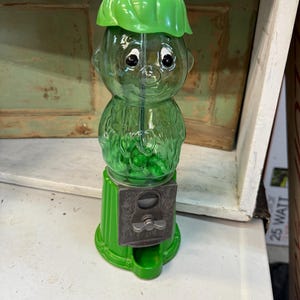 May include: A vintage green glass gumball machine shaped like a whimsical character with a green hat. The machine has a silver coin slot and a dispensing area. The character's face has black eyes and a smiling expression. The base is a bright green color.