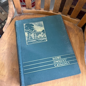 May include: A dark green hardcover book titled Home Owners' Catalogs with gold lettering and a gold-embossed illustration of a house and trees on the front cover. The text Home Sweet Home is printed below the illustration on the front cover.