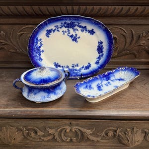 May include: A collection of antique blue and white porcelain serving dishes. The set includes a large oval platter, a covered serving bowl, and a small oblong dish. The pieces feature floral patterns and gold trim.