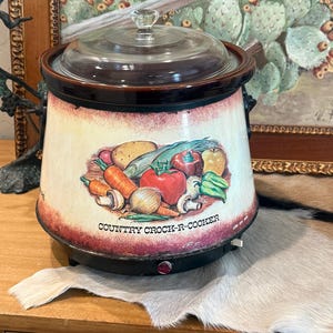 May include: A vintage Country Crock-R-Cooker slow cooker with a glass lid. The cream-colored crock features a colorful illustration of vegetables. The base is black with a red power indicator light. The text "COUNTRY CROCK-R-COOKER" is printed on the front.
