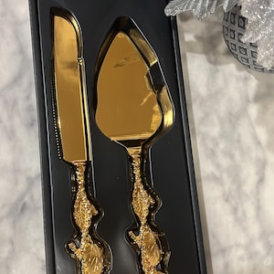 Puede incluir: Un juego de servir pasteles de color dorado que consta de un cuchillo dentado y una pala triangular. Ambos utensilios cuentan con mangos moldeados con un diseño tridimensional de rosas y hojas, presentados en una caja negra.