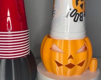 Puede incluir: Dos portavasos con temática de Halloween, uno negro y uno naranja. El portavasos negro tiene una pila de vasos rojos en la parte superior. El portavasos naranja tiene forma de calabaza con un vaso blanco en su interior. Dos velas de té blancas están sobre la mesa.