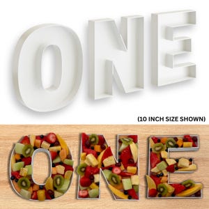 Puede incluir: Letras blancas tridimensionales que deletrean "ONE" llenas de fruta colorida. Las letras se muestran sobre una superficie de madera. También es visible el texto "(10 INCH SIZE SHOWN)".