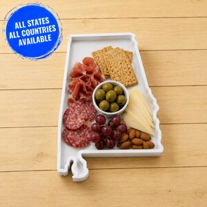 Könnte beinhalten: Ein weißes Serviertablett in Form des Bundesstaates Alabama mit einer kleinen weißen Schale mit grünen Oliven, Crackern, Salami, Prosciutto, Käseecken und Mandeln. Ein blaues rundes Abzeichen zeigt den Text ALL STATES ALL COUNTRIES AVAILABLE.