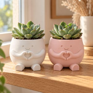 May include: Two round ceramic planters, one white and one light pink, each featuring a sculpted face and arms forming a heart shape. Each planter has two small feet and contains a green succulent plant with pointed, fleshy leaves.