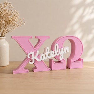 Könnte beinhalten: Ein rosafarbenes Chi-Omega-Sorority-Schild mit den griechischen Buchstaben Chi und Omega in matter Ausführung. Der in weißer Schreibschrift gehaltene Name Katelyn ist mittig über die rosa griechischen Buchstaben gelegt und erzeugt einen 3D-Effekt.