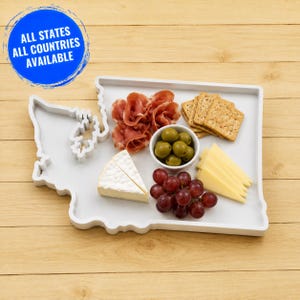 May include: A white serving tray shaped like the state of Washington, featuring a blue circular badge with the text ALL STATES ALL COUNTRIES AVAILABLE. The tray holds slices of brie, prosciutto, green olives in a bowl, grapes, and crackers.