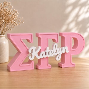 Peut inclure: Un signe tridimensionnel en lettres grecques roses représentant les caractères Sigma, Gamma et Rho. Une plaque nominative blanche en écriture cursive avec le texte Katelyn est superposée au centre des lettres roses en forme de blocs.