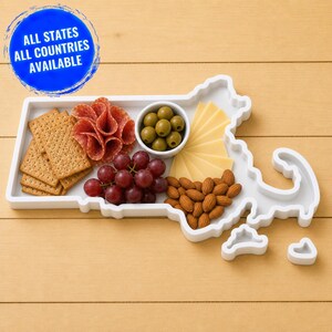 Könnte beinhalten: Ein weißes Serviertablett in Form des Bundesstaates Massachusetts, das eine kleine weiße Schale mit grünen Oliven, Cracker, gefaltete Salamischeiben, rote Weintrauben und Mandeln enthält. Eine blaue kreisförmige Grafik zeigt den Text ALL STATES ALL COUNTRIES AVAILABLE.