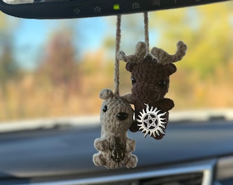 May include: Two crocheted car charms hanging from a rearview mirror. One is a light brown bear with a bronze-colored pendant. The other is a brown moose with a silver-colored pentacle charm. Both are suspended by beige strings.