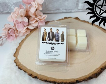 May include: A clear plastic package containing six square wax melts. The package has a label with the word "SUPERNATURAL" and images of four people. The wax melts are light yellow and sit on a wooden slice with pink flowers.