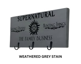 May include: A weathered gray wooden key holder with three hooks. The key holder features the text "Supernatural", "Saving People", "Hunting Things", "The Family Business", and a black and white symbol.