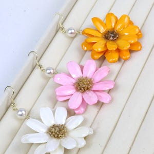 May include: Three pairs of dangle earrings featuring flower-shaped charms in orange, pink, and white. Each earring has a gold-tone metal hook, a single white faux pearl bead, and a textured center on the flower petals with a glossy finish.